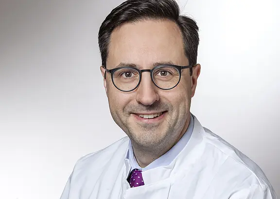 Porträt eines mittelalten Mannes mit Brille im weißen Arztkittel. Er lächelt freundlich in die Kamera; violette Krawatte sichtbar. Neutraler, heller Hintergrund.