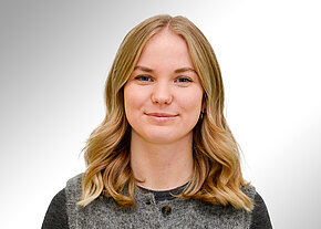 Denise Schellmann Porträtfoto: Junge Frau mit schulterlangen, leicht welligen blonden Haaren blickt in die Kamera und lächelt dezent. Sie trägt ein graues Oberteil. Heller, neutraler Hintergrund.