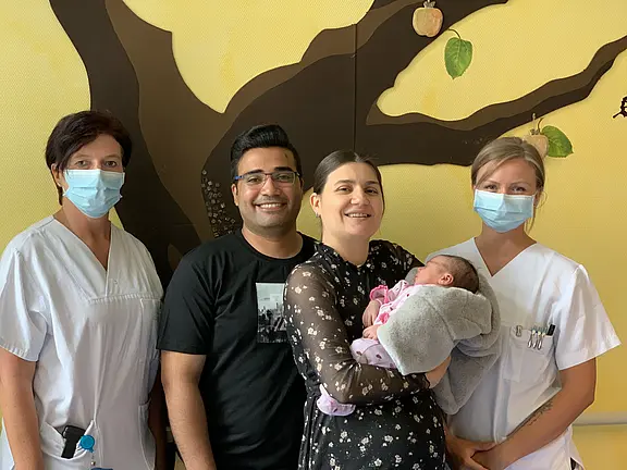 Vier Personen stehen in einem Raum mit einer Wanddekoration eines Baumes. Eine Frau hält ein neugeborenes Baby in einem grauen Tuch, umgeben von zwei medizinischen Fachkräften und einem Mann. Alle tragen Mund-Nasen-Schutz. Die Stimmung wirkt freundlich und feierlich.