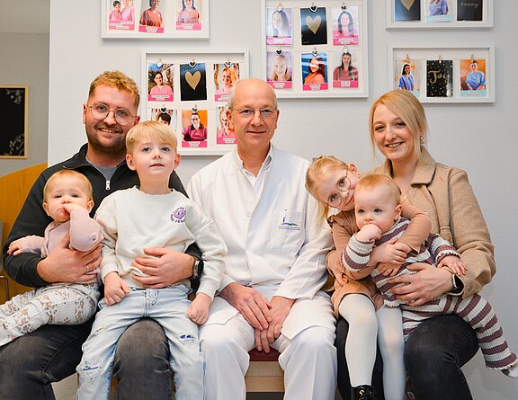 Eine Familie mit drei kleinen Kindern sitzt in einer Arztpraxis. Vater links hält ein Baby, der Sohn sitzt auf seinem Schoß, in der Mitte ein älterer Arzt im weißen Kittel, rechts die Mutter mit einem Baby. Alle lächeln freundlich in die Kamera.