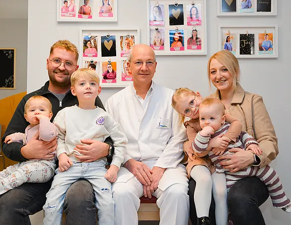 Eine Familie mit drei kleinen Kindern sitzt in einer Arztpraxis. Vater links hält ein Baby, der Sohn sitzt auf seinem Schoß, in der Mitte ein älterer Arzt im weißen Kittel, rechts die Mutter mit einem Baby. Alle lächeln freundlich in die Kamera.