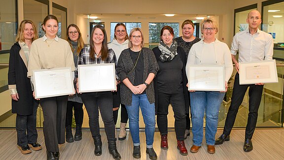 Eine Gruppe von acht Personen steht in einem modernen Innenraum. Sie halten Dokumente oder Urkunden in den Händen und lächeln. Im Hintergrund sind große Fenster sichtbar, die Tageslicht hereinlassen. Es wirkt wie eine Preisverleihung oder Anerkennungsveranstaltung.