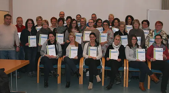 Gruppenfoto von rund 30 Erwachsenen in einem Raum. Viele sitzen vorne, andere stehen dahinter. Alle halten Zertifikate in den Händen und lächeln. Es wirkt wie der Abschluss oder die Verleihung von Teilnahmeurkunden eines Kurses oder einer Schulung.