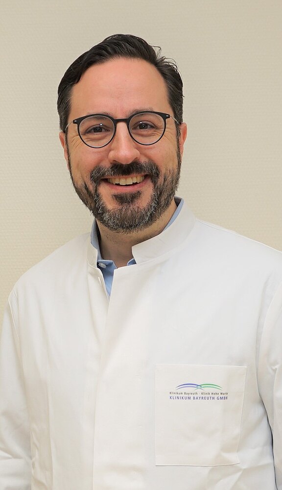 Lächelnder Arzt mit Brille im weißen Kittel steht vor neutraler Wand, wirkt freundlich und professionell.