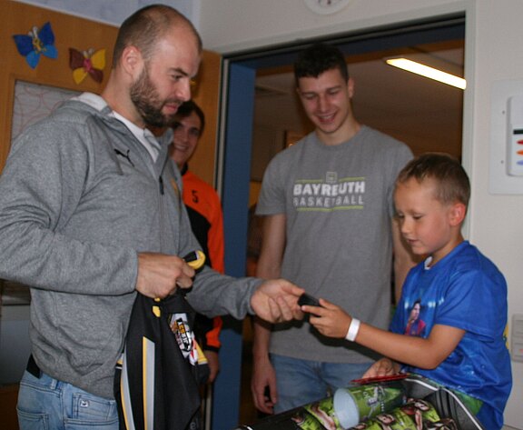 Mehrere Basketballspieler besuchen einen Jungen im Krankenhaus. Einer überreicht dem Kind ein kleines Geschenk, während andere lächelnd zusehen. Auf dem Bett liegen Fanartikel und ein Trikot. Die Szene zeigt einen freundlichen Austausch und Unterstützung für den jungen Patienten.