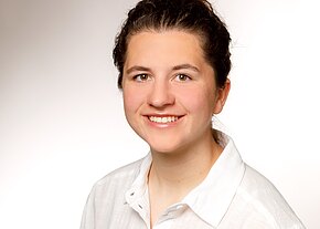 Professionelles Porträt einer freundlich lächelnden jungen Frau mit hochgesteckten Haaren in weißem Hemd vor neutral hellem Hintergrund.