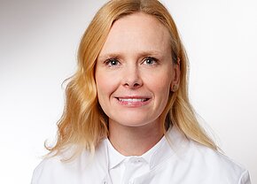 PD Dr. med. Anne Marie Augustin Ein Portrait einer Frau mit blonden, lockigen Haaren, die in einem weißen Arztkittel gekleidet ist. Sie hat ein freundliches Lächeln und schaut direkt in die Kamera. Der Hintergrund ist hell und einheitlich.