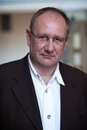 Porträt eines mittelalten Mannes mit Brille. Er trägt ein dunkles Sakko über einem weißen Hemd und blickt ernst in die Kamera. Neutraler, unscharfer Hintergrund.