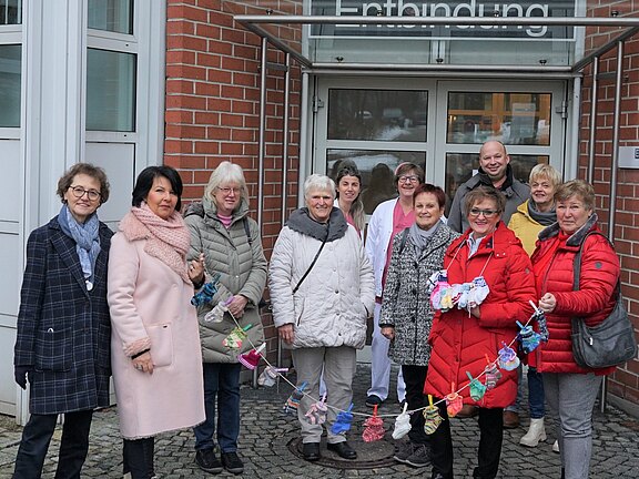 Vor dem Eingang der Entbindungsstation steht eine Gruppe, darunter Krankenhauspersonal. Sie halten eine lange Girlande mit gestrickten Babysocken und Mützen – eine freundliche Geste oder Spende für Neugeborene.
