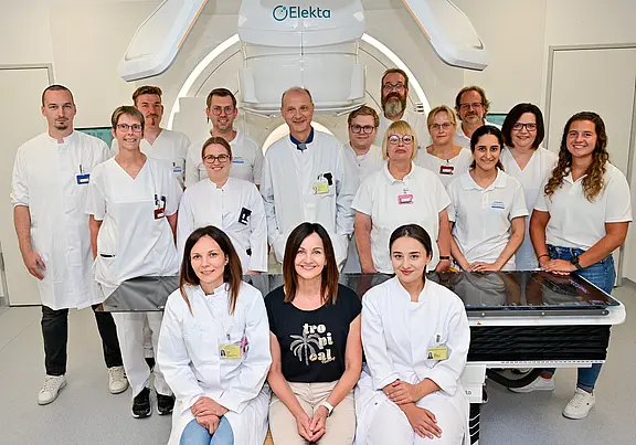 Gruppenfoto eines medizinischen Teams vor einem großen Strahlentherapiegerät in einer Klinik. Die meisten tragen weiße Kittel; zwei Personen in Alltagskleidung sitzen in der ersten Reihe, flankiert von Mitarbeitenden.