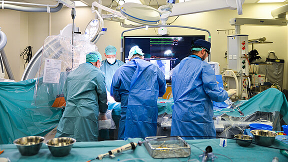 Im Bild sind vier Chirurgen in einem Operationssaal zu sehen, die an einem OP-Tisch arbeiten. Sie tragen Schutzkleidung und Mundschutz. Ein Monitor im Hintergrund zeigt medizinische Daten. Der Raum ist mit verschiedenen medizinischen Geräten ausgestattet.