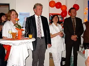 Eine Gruppe von fünf Personen steht in einem festlich dekorierten Raum. Sie tragen elegante Kleidung und lächeln. An einem Stehtisch stehen Getränke. Im Hintergrund sind rote Luftballons sichtbar, was auf eine Feier hinweist.