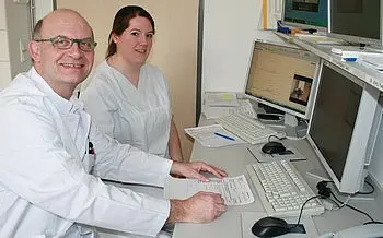 Ein Arzt und eine Pflegekraft sitzen an einem Schreibtisch in einem Büro. Beide tragen weiße Kittel und lächeln. Auf dem Tisch sind Computer und Dokumente sichtbar, während der Arzt einen Arbeitsablauf oder ein Formular ausfüllt.