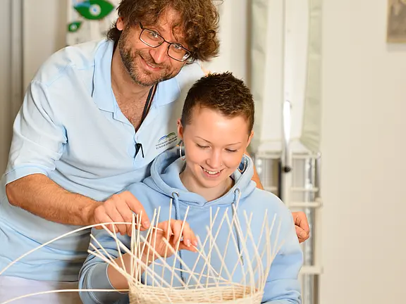 Ein erwachsener Betreuer zeigt einer jungen Person das Korbflechten; beide lächeln, und ihre Hände formen gemeinsam ein Geflecht aus hellen Ruten.