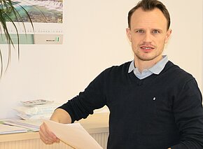 Florian Bär Ein Mann sitzt an einem Tisch und hält ein Blatt Papier in der Hand. Er trägt einen dunkelblauen Pullover mit einem hellblauen Kragen. Der Hintergrund zeigt eine Wand mit einem Kalender und einer Pflanze.