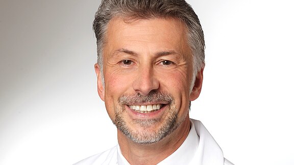 Ein lächelnder Mann mit grauen Haaren und einem Vollbart trägt ein weißes Hemd. Der Hintergrund ist hell und neutral. Der Ausdruck ist freundlich und einladend.