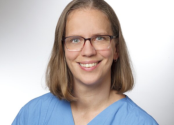 Porträt einer lächelnden Person mit schulterlangem Haar und Brille, gekleidet in blauer medizinischer Kleidung, vor hellem, neutralem Hintergrund.