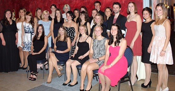 Eine Gruppe von etwa 30 Personen posiert zusammen in einem festlichen Raum. Die Frauen tragen elegante Kleider in verschiedenen Stilen und Farben, während ein paar Männer in Anzügen stehen und sitzen. Die Atmosphäre wirkt freundlich und feierlich.