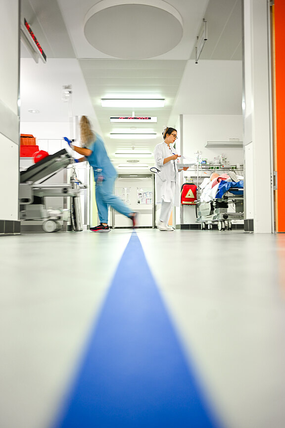Heller Krankenhausflur mit blauer Bodenlinie, die in einen Behandlungsraum führt. Pflegekräfte arbeiten; eine schiebt eine Trage, eine liest Unterlagen. Eindruck von Notfallbereich und Bewegung.