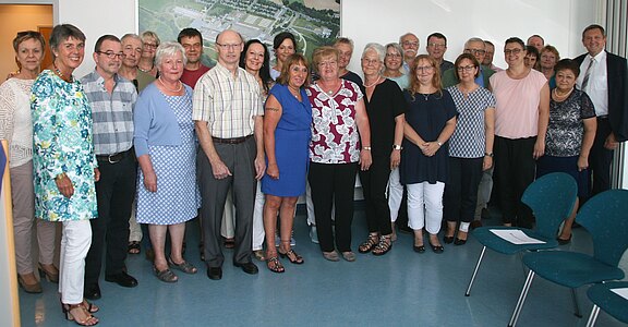 Gruppe von rund 25 Erwachsenen steht dicht zusammen in einem hellen Raum und blickt lächelnd in die Kamera. Mischungen aus Freizeit- und Businesskleidung, sommerlich. Es wirkt wie ein Gruppenfoto bei einem Treffen oder einer Feier.