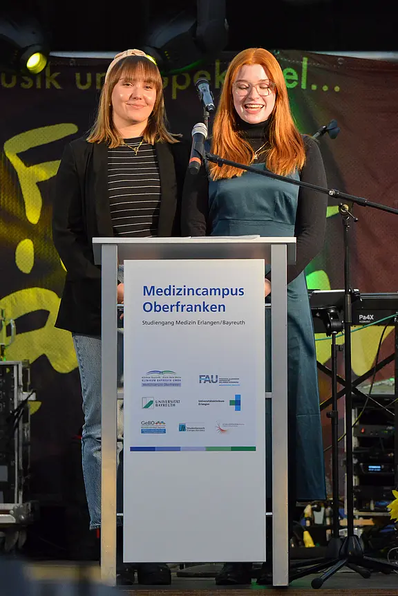 Zwei Frauen stehen an einem Rednerpult und lächeln ins Publikum. Das Pult zeigt das Logo und den Namen „Medizincampus Oberfranken“. Im Hintergrund sind unscharfe musikalische Elemente zu erkennen. Die Veranstaltung scheint feierlich und informativ zu sein.