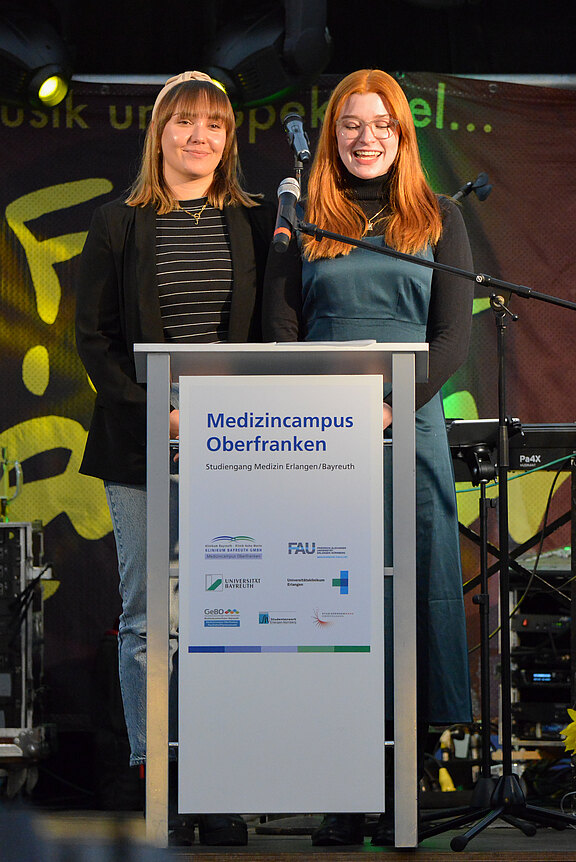Zwei Frauen stehen an einem Rednerpult und lächeln ins Publikum. Das Pult zeigt das Logo und den Namen „Medizincampus Oberfranken“. Im Hintergrund sind unscharfe musikalische Elemente zu erkennen. Die Veranstaltung scheint feierlich und informativ zu sein.