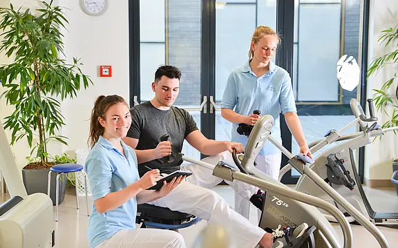 Reha-Training in heller Praxis: Ein junger Mann trainiert an einem Kardio-/Bewegungsgerät, begleitet von zwei Therapeutinnen in hellblauer Kleidung. Eine notiert Werte auf einem Tablet, die andere stellt das Gerät ein.