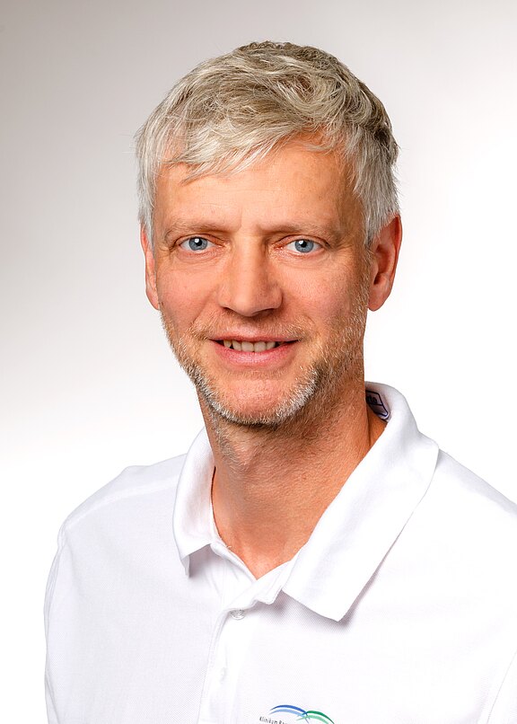 Ein lächelnder Mann mit kurzen, grauen Haaren und blauen Augen trägt ein weißes Polohemd mit einem Logo. Der Hintergrund ist hell und einfarbig, was den Fokus auf sein Gesicht lenkt.