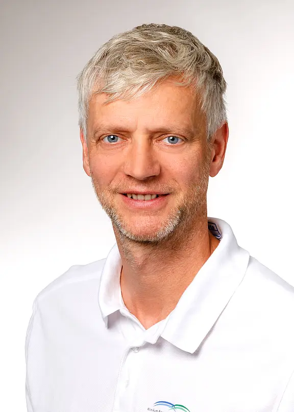 Ein lächelnder Mann mit kurzen, grauen Haaren und blauen Augen trägt ein weißes Polohemd mit einem Logo. Der Hintergrund ist hell und einfarbig, was den Fokus auf sein Gesicht lenkt.