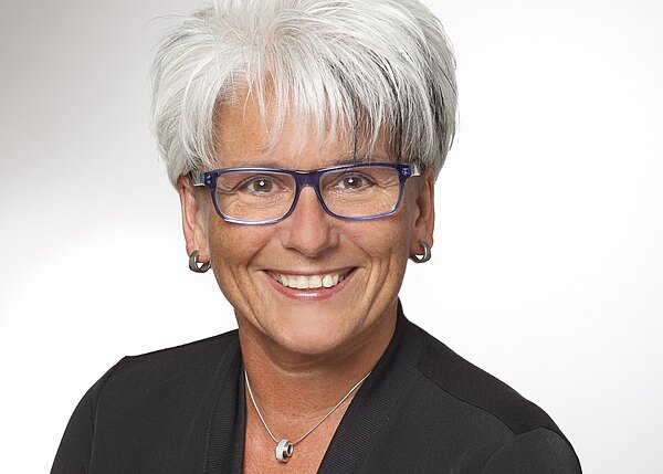 Porträt einer lächelnden Frau mit kurzen, grauweißen Haaren und blauer Brille. Sie trägt einen dunklen Blazer, kleine Ohrringe und eine silberne Kette. Neutraler, heller Hintergrund; Fokus auf Gesicht und freundlichem Ausdruck.