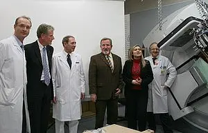 Sechs Personen in weißen Kitteln und formeller Kleidung stehen in einem Raum. Sie lächeln und scheinen ein Gespräch zu führen, während im Hintergrund ein medizinisches Gerät zu sehen ist. Die Atmosphäre ist feierlich und kollegial.