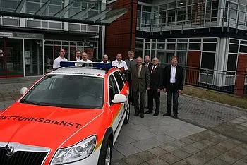 Eine Gruppe von Menschen steht vor einem Einsatzfahrzeug des Rettungsdienstes. Das Auto ist orange-weiß lackiert und trägt Licht- und Schriftzeichen. Im Hintergrund ist ein modernes Gebäude sichtbar, das in ein Programm für Rettungsdienste integriert ist.