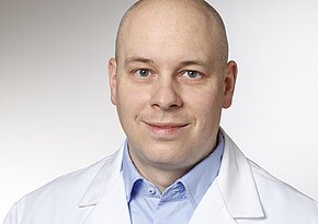 Sven-Torge Brandt Ein mittelalter Mann mit Glatze trägt einen weißen Arztkittel über einem blauen Hemd. Er lächelt in die Kamera und hat blaue Augen. Der Hintergrund ist hell und neutral gehalten.