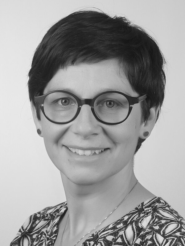 Schwarz-weißes Porträt einer lächelnden Person mit kurzen Haaren und runder Brille, die frontal in die Kamera blickt; neutraler Hintergrund.