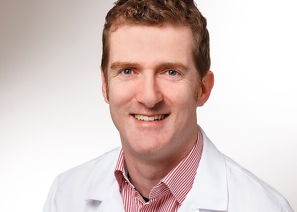 Porträt einer lächelnden Person in weißem Kittel vor hellem Hintergrund, mit Blick in die Kamera. Unter dem Kittel ein rot-weiß gestreiftes Hemd.