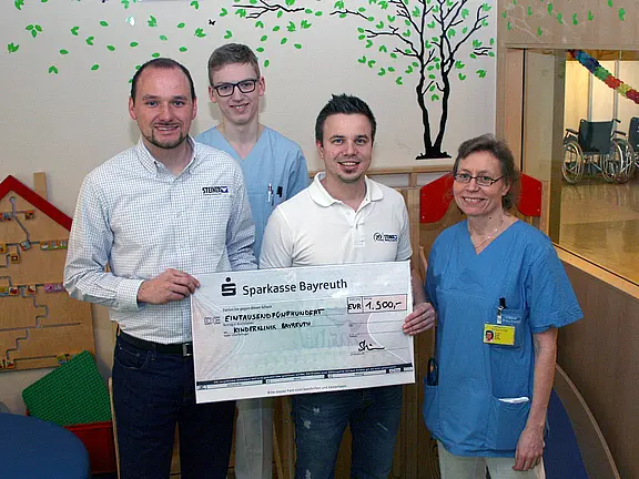 Vier Erwachsene, zwei in Klinikbekleidung, posieren in einem Krankenhaus und halten einen übergroßen Spendenscheck der Sparkasse Bayreuth über 1.500 Euro. Gruppenfoto zur Spendenübergabe.