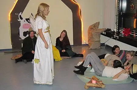 Eine Gruppe von Menschen sitzt auf dem Boden in einem Raum. Eine Person in einem weißen Kleid steht im Vordergrund und spricht. Im Hintergrund sind einige dekorative Elemente zu sehen. Die anderen Zuhörer scheinen aufmerksam zu sein und verfolgen die Darbietung.