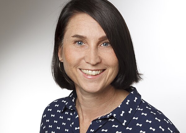 Porträt einer lächelnden Frau mit schulterlangem, dunkelbraunem Haar. Sie trägt ein blaues Hemd mit weißen Schleifenmustern. Der Hintergrund ist hell und unifarben. Ihr Gesichtsausdruck wirkt freundlich und einladend.