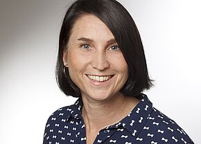 Verena Brüls Porträt einer lächelnden Frau mit schulterlangem, dunkelbraunem Haar. Sie trägt ein blaues Hemd mit weißen Schleifenmustern. Der Hintergrund ist hell und unifarben. Ihr Gesichtsausdruck wirkt freundlich und einladend.