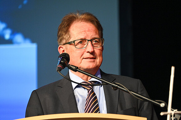 Mann mit Brille in Anzug und gestreifter Krawatte spricht am Rednerpult in einem Saal. Er steht hinter einem Mikrofon, Blick nach vorn. Bühne und neutraler Hintergrund, Fokus auf die Rede.