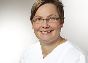Silke Friedrich Porträt einer lächelnden Person mit kurzer Frisur und Brille in weißer medizinischer Arbeitskleidung, vor neutralem Hintergrund, direkt in die Kamera blickend.
