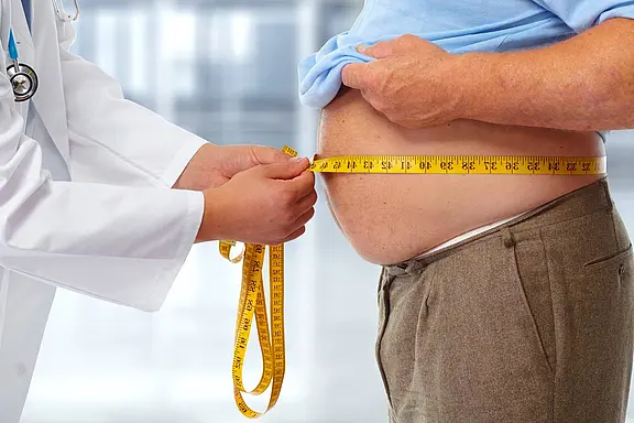 Ein Arzt misst den Bauchumfang eines Patienten mit einem gelben Maßband. Der Patient trägt ein hellblaues Hemd und eine dunkle Hose. Der Hintergrund ist unscharf und scheint eine medizinische Umgebung zu sein.