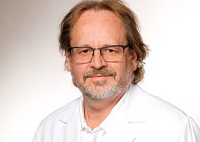 Frontales Porträt eines mittelalten Mannes mit Brille, kurzen welligen Haaren und grauem Bart. Er trägt einen weißen Kittel und Hemd und blickt ruhig in die Kamera vor hellem, neutralem Hintergrund.