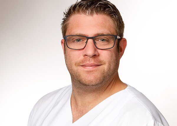 Porträt eines Mannes mit Brille in weißer medizinischer Kleidung vor hellem Hintergrund; er blickt freundlich in die Kamera.