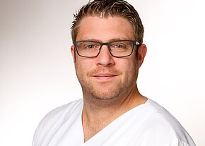 Station 23 Porträt eines Mannes mit Brille in weißer medizinischer Kleidung vor hellem Hintergrund; er blickt freundlich in die Kamera.