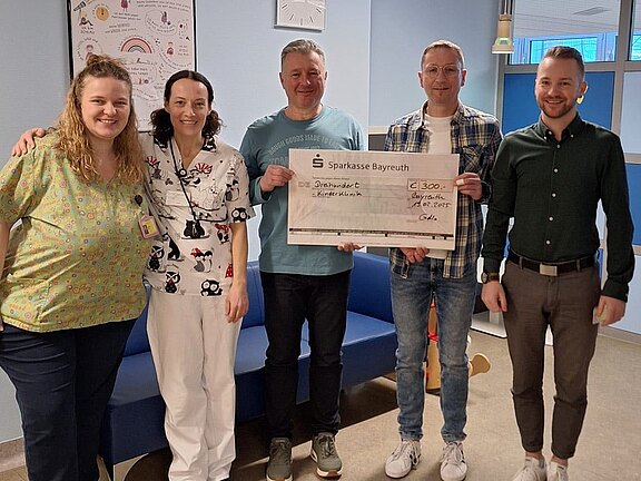 Fünf Erwachsene in einem Klinikflur lächeln; zwei in der Mitte halten einen großen Spendenscheck der Sparkasse Bayreuth über 1.500 Euro.