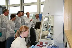In einem Labor arbeitet eine Frau am Tisch, während mehrere Personen in weißen Laborkitteln von ihr lernen. Die Gruppe schaut aufmerksam zu, während sie an einem Experiment beteiligt ist.