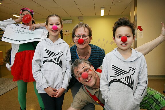 Vier Personen, darunter zwei Kinder, tragen rote Clownnasen und lächeln in die Kamera. Im Hintergrund hält eine verkleidete Person einen großen Spendenscheck. Die Szene wirkt fröhlich und gemeinnützig, als Teil einer Aktion von Klinikclowns oder einer Benefizveranstaltung.