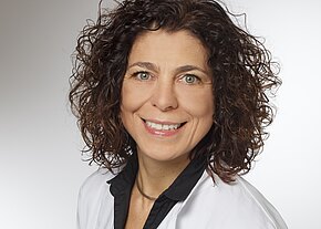 Sabine Tröger Porträt einer lächelnden Frau mit lockigem braunem Haar, in weißem Kittel und dunklem Kragen, vor neutralem Hintergrund.