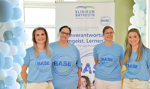 Vier Personen in hellblauen BASE-T-Shirts posieren vor einem Banner des Klinikums Bayreuth. Die Szene wirbt für die Ausbildungsstation BASE; freundliches Gruppenfoto bei einer Informations- oder Eröffnungsveranstaltung.
